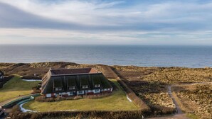 A coastal building with a sloped roof, surrounded by a grassy area and a rocky shoreline. - Lovingly designed 62m² apartment offers space for 3 people on the 1st floor. - (Kampen (Sylt))