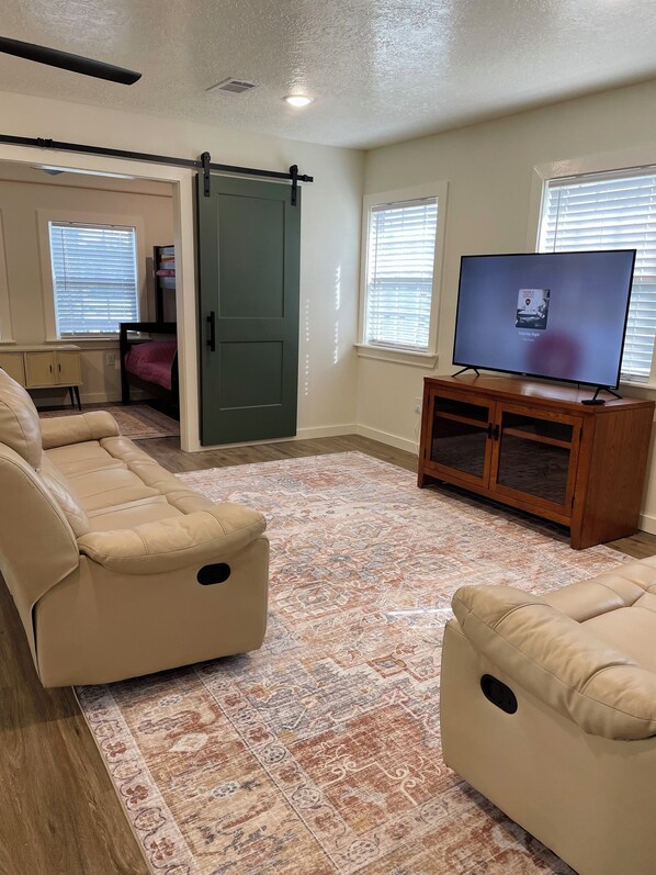 A living room with a large flat-screen TV, beige leather sofas, a wooden entertainment unit, and a patterned rug. - Under the Pines on Lake Livingston- BEAUTIFUL WATER VIEW! (Onalaska)