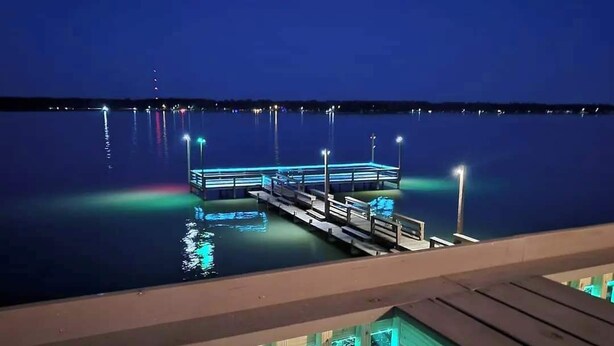 Ein Steg, der in ruhiges Wasser in der Nacht hineinreicht, wobei sich Lichter auf der Oberfläche spiegeln.