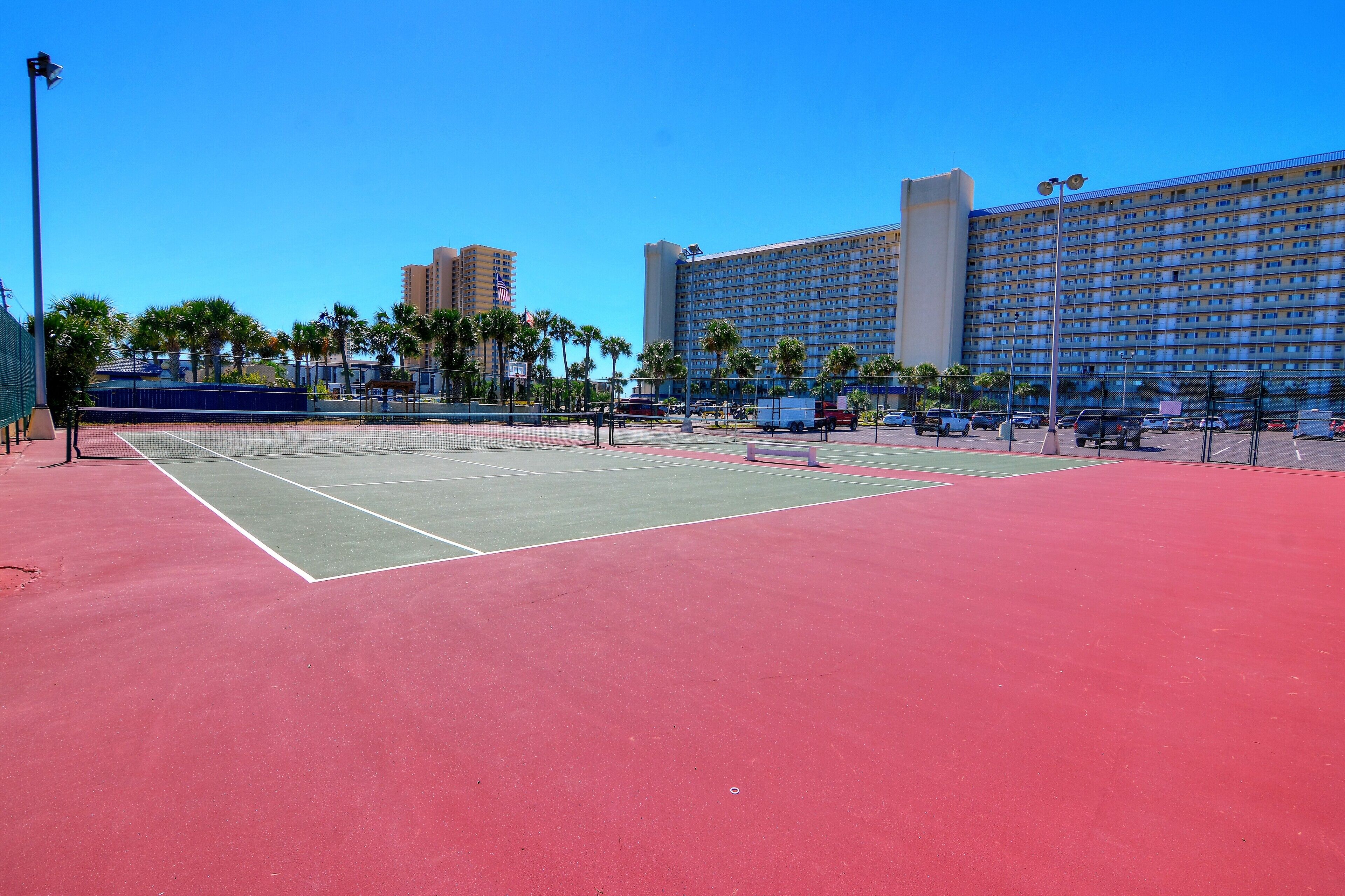 A tennis court with a red surface, surrounded by a fence, and a large hotel building in the background.