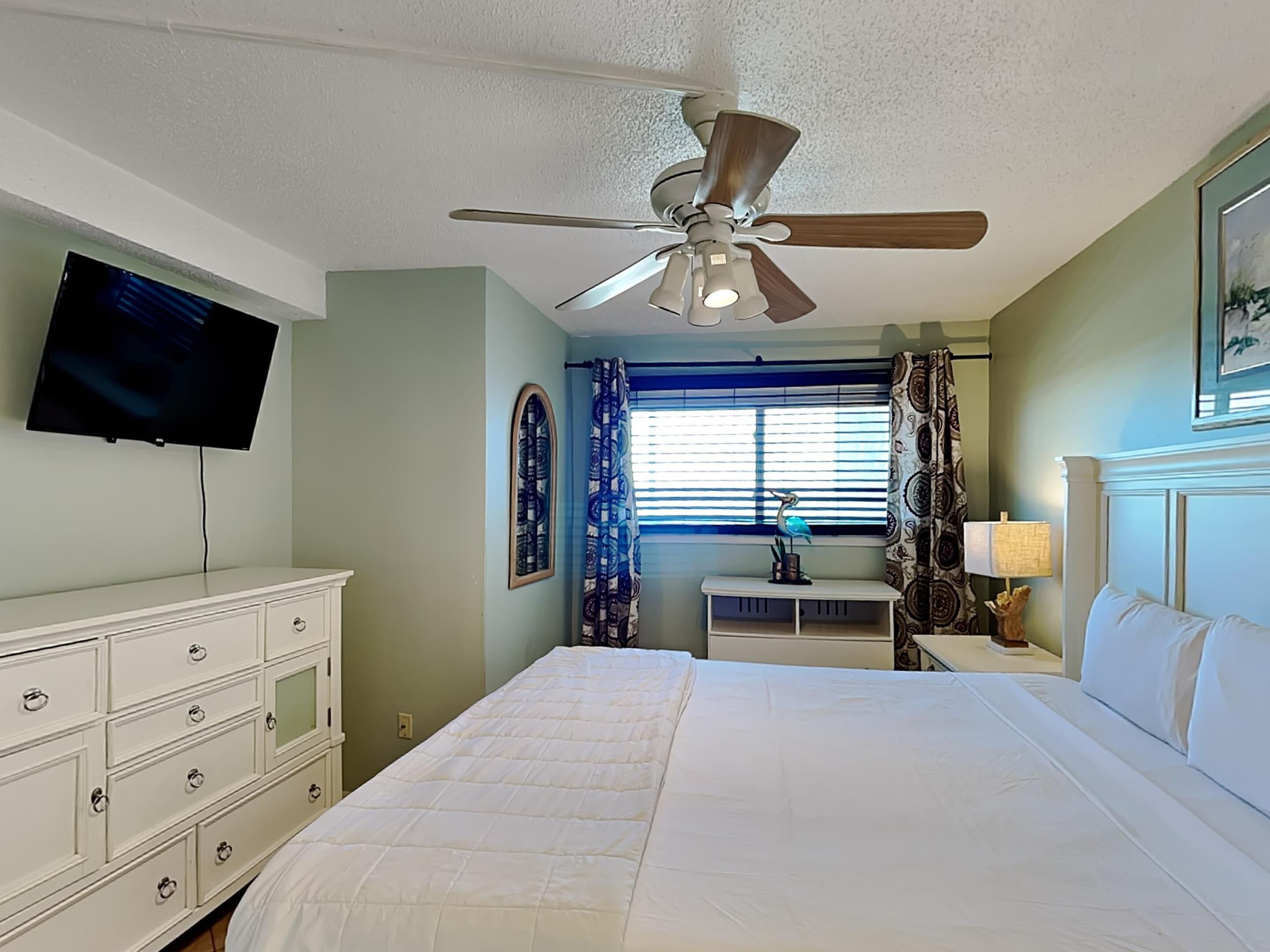 A bedroom with a bed, a ceiling fan, a TV mounted on the wall, a desk with a lamp, and a window with curtains.