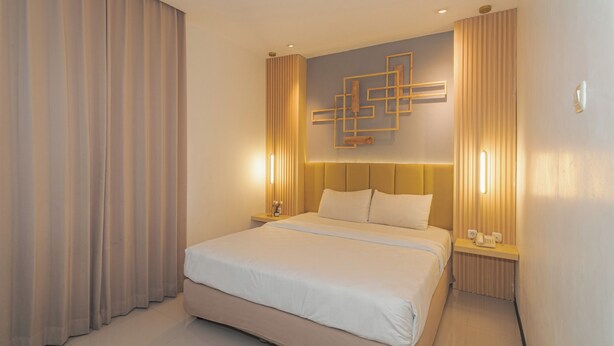 A hotel room with a large bed, a headboard with integrated lighting, a bedside table with a lamp, and a wall-mounted artwork. - Best Hotel (Surabaya)