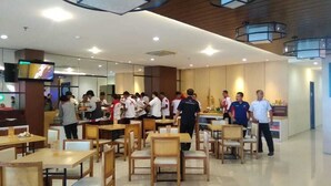 A spacious dining area with wooden tables and chairs, a television mounted on a pillar, and a modern ceiling design. - Best Hotel (Surabaya)