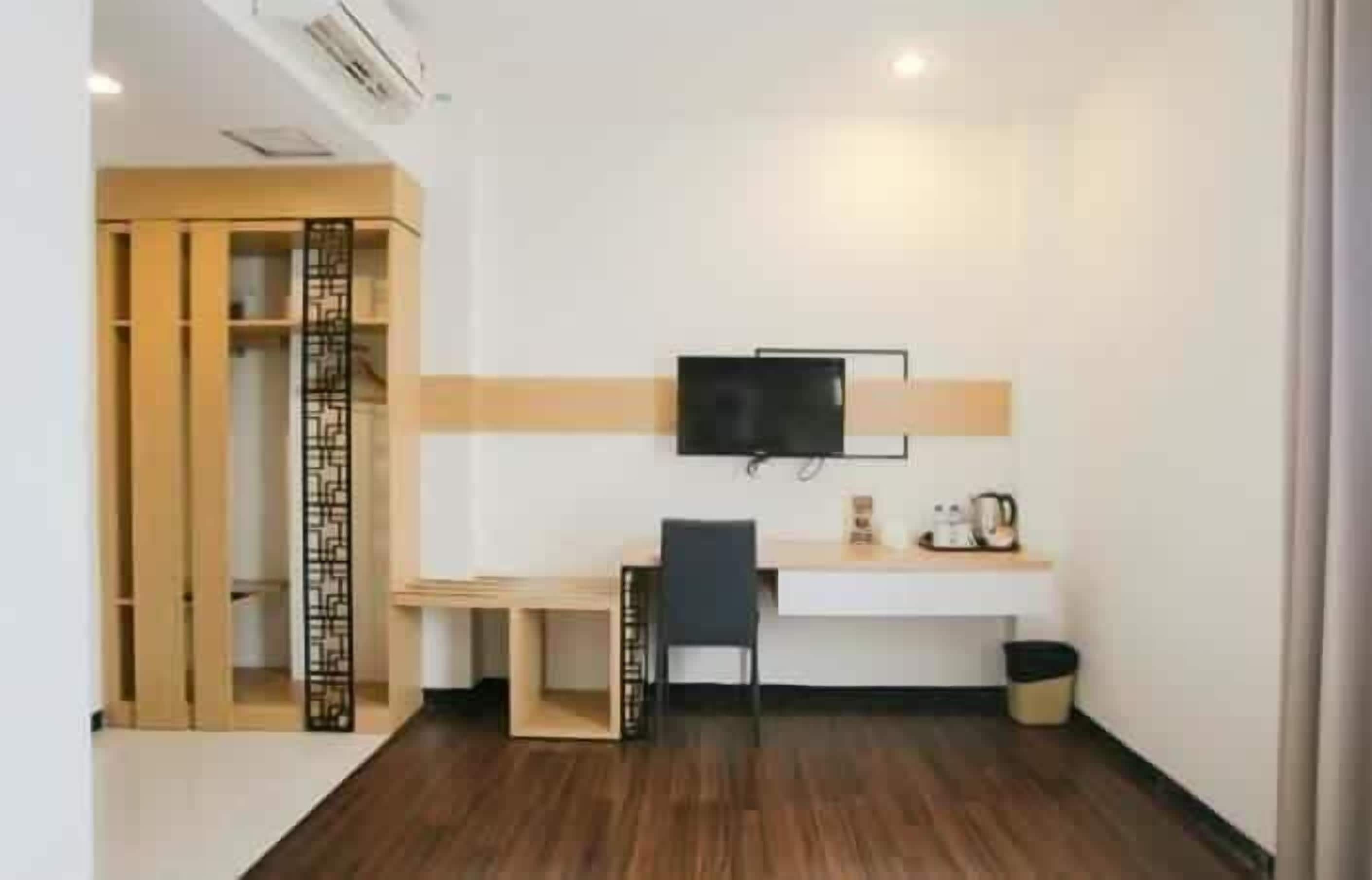 A compact hotel room with a wooden floor, a desk with a chair, a television mounted on the wall, and a small shelf with a kettle and other items.
