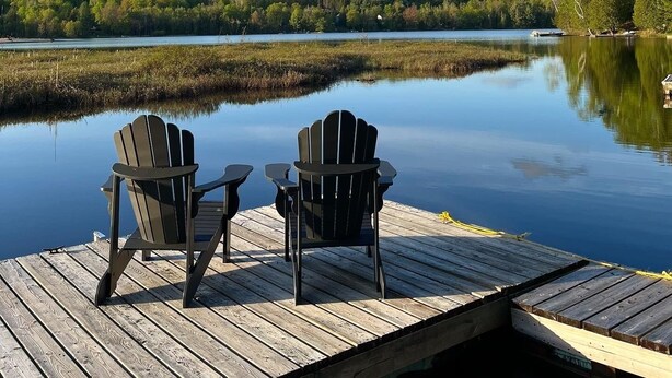 Puinen laituri, jossa on kaksi Adirondack-tyylistä tuolia, ja ne on asetettu niin, että ne osoittavat rauhallista järveä kohti. Laiturin ympärillä on puita ja taivaalla ei ole pilviä.
