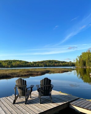 A wooden dock with two Adirondack chairs facing a calm lake, surrounded by trees and a clear sky.
