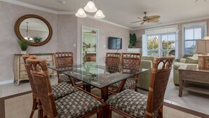 A dining area with a glass-top table surrounded by wicker chairs with patterned cushions, a ceiling fan, and a large window with blinds.