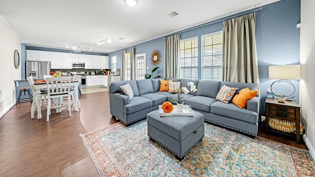 A modern living room with a grey sectional sofa, a dining table with chairs, and a kitchen in the background.