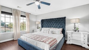 A bedroom with a large bed, a ceiling fan, two bedside tables with lamps, and a view of the outdoors through a window.