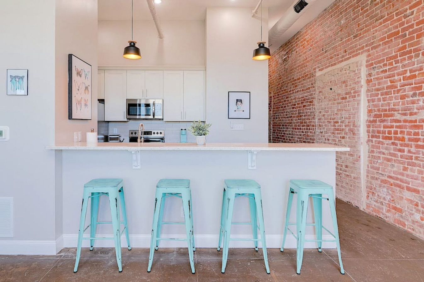 A modern kitchen with white cabinets, a brick wall, and three light blue bar stools.