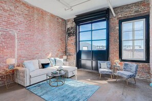 Ein geräumiges Wohnzimmer mit freiliegenden Ziegelwänden, einem großen Fenster, einem weißen Sofa, einem kleinen runden Couchtisch und zwei gestreiften Sesseln.