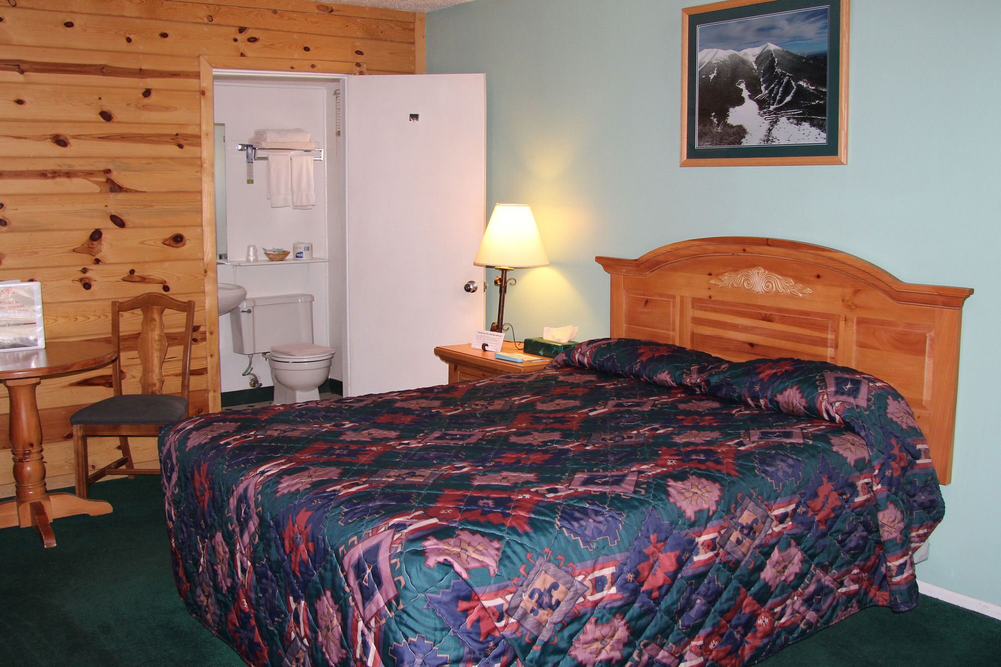 A hotel room with a wooden headboard, a bed with a patterned bedspread, a wooden chair, a small table with a lamp, and a bathroom visible through an open door.