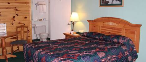 A hotel room with a wooden headboard, a bed with a patterned bedspread, a wooden chair, a small table with a lamp, and a bathroom visible through an open door.