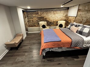 A bedroom with a stone wall, a bed with multiple pillows, a bench, and a small table.