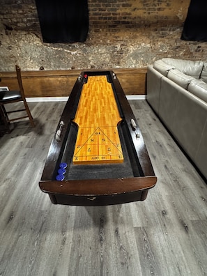 A shuffleboard table with a wooden playing surface and blue pucks, set against a brick wall and a leather sofa. - Carrollton Square's exclusive VRBO Suite. (Carrollton)