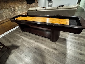 A billiards table with a wooden playing surface and a dark frame, set on a wooden floor in a room with a brick wall and a white couch.