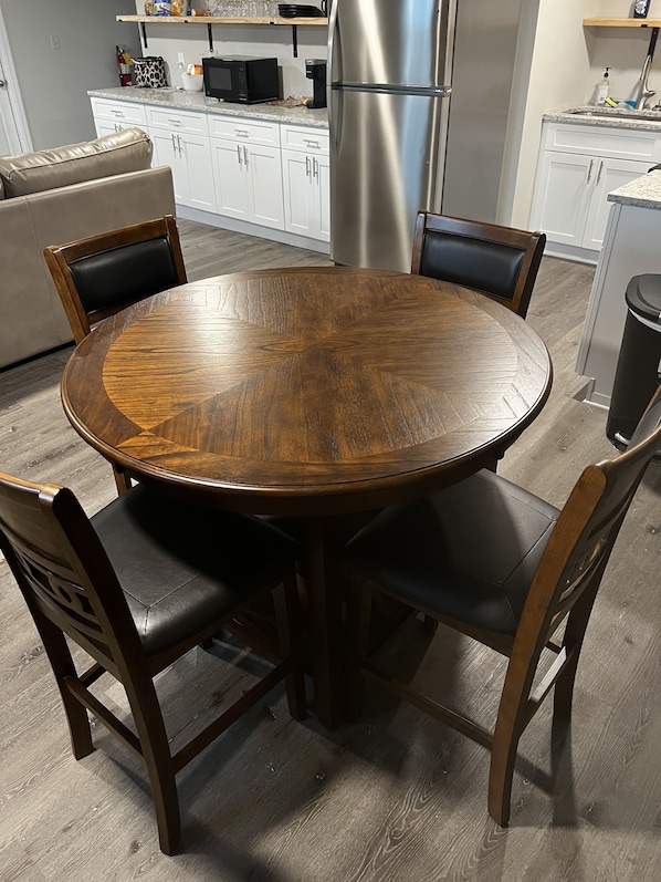 A round wooden dining table with four chairs, a kitchen area with white cabinets, a stainless steel refrigerator, and a countertop with various items.