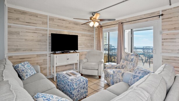 A living room with a large sofa, a television on a white cabinet, and a ceiling fan. - Oceanfront Condo-large, corner balcony, enclosed pool in popular Cherry Grove Beach (North Myrtle Beach)