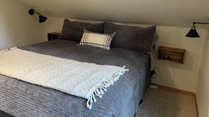Un lit avec une couette grise, un plaid blanc et deux oreillers. Il y a deux tables de chevet avec des lampes.