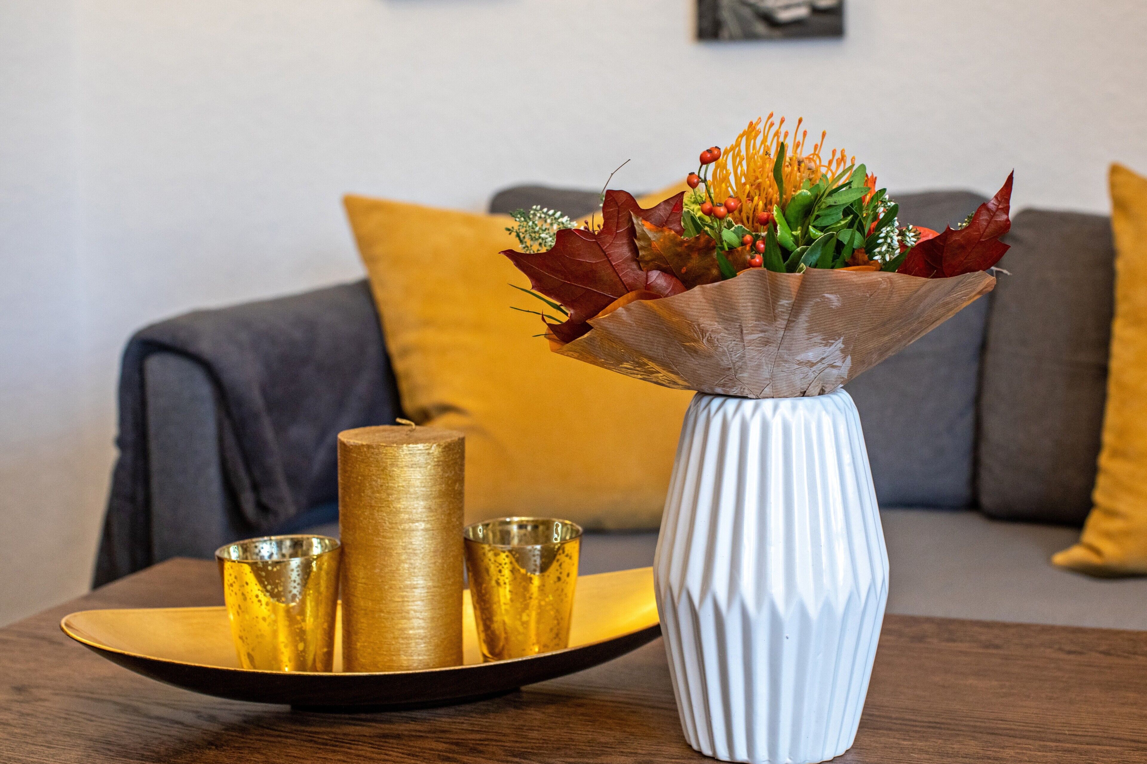 A white vase with a textured design, a golden candle, and a golden cup on a tray, placed on a wooden table in a room with a grey sofa and a yellow cushion.