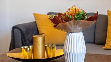 A white vase with a textured design, a golden candle, and a golden cup on a tray, placed on a wooden table in a room with a grey sofa and a yellow cushion.