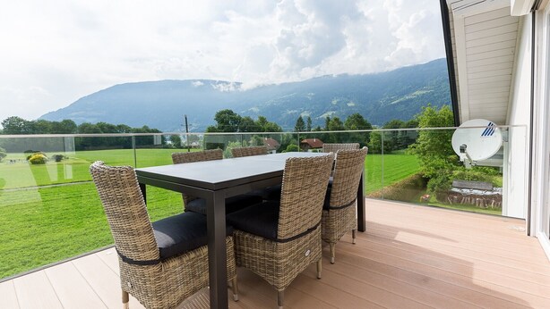 Een balkon met een tafel, stoelen en een glazen balustrade, met uitzicht op bergen.