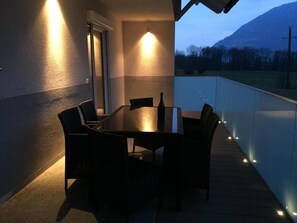 Un área de comedor al aire libre con una mesa, sillas, barandilla de cristal y vistas a la montaña al fondo.