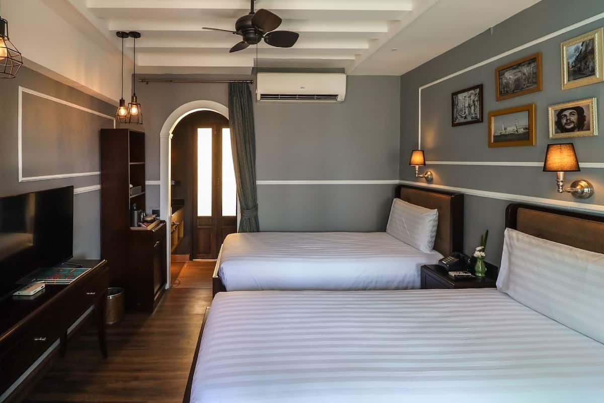 Hotelkamer met twee bedden, een televisie, een bureau en een kledingkast. Er zijn wandlampen en een plafondventilator.