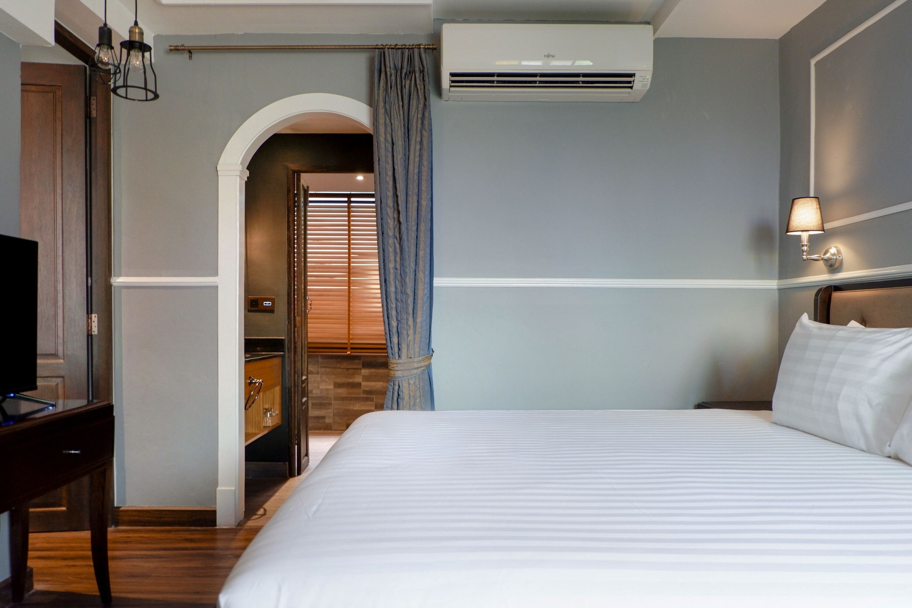 Une chambre d’hôtel avec un grand lit, un bureau avec une télévision, un appareil de climatisation et une salle de bain visible à travers une arche.