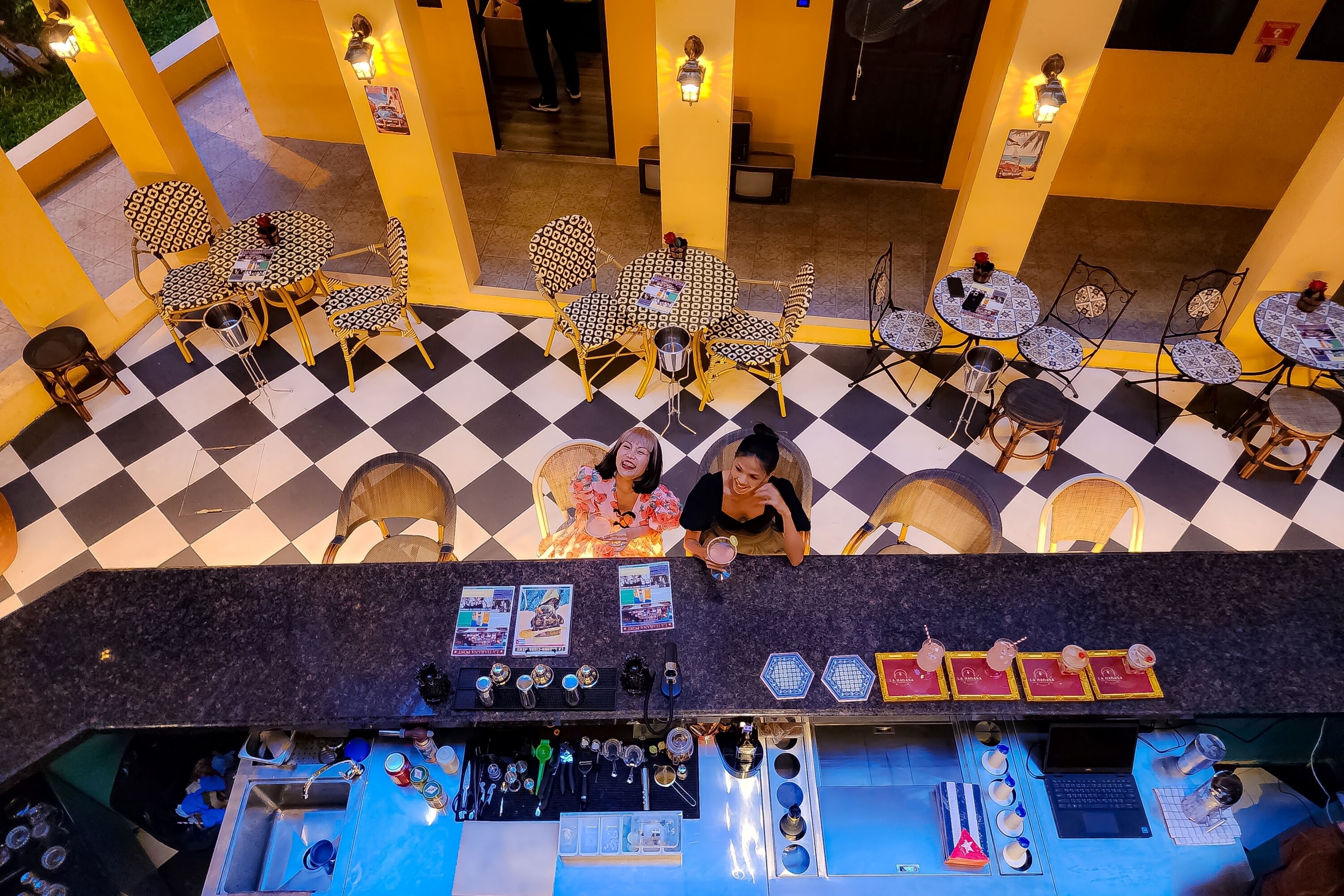A bar area with checkered flooring, patterned chairs, and a counter with various bar tools and glasses.