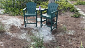 Dos sillas Adirondack verdes con una mesita y un florero, ubicadas en un sendero de piedra rodeado de vegetación y un edificio al fondo.
