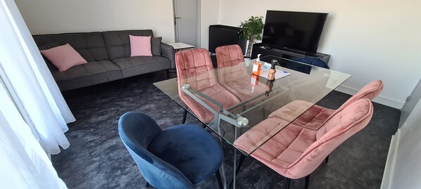 A modern living room with a glass-top dining table, two pink cushioned chairs, a blue cushioned chair, a grey sofa with pink cushions, a black TV stand, and a potted plant. - Apartment 65m² Paris ♡ Disney ♡ Centrex ☆ RER A (Noisy-le-Grand)
