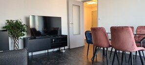 A modern living room with a flat-screen TV, a black entertainment unit, a potted plant, and a dining area with pink and blue chairs. - Apartment 65m² Paris ♡ Disney ♡ Centrex ☆ RER A (Noisy-le-Grand)