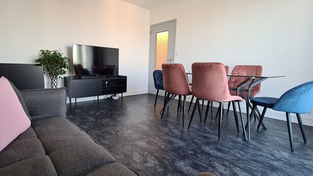 Un salon moderne avec un canapé gris, un téléviseur, une table à manger avec des chaises et une plante.