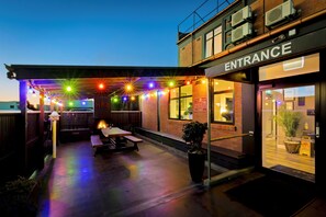 Un espace extérieur aménagé en terrasse, avec des guirlandes lumineuses, une plateforme en bois et l’entrée d’un bâtiment en briques.