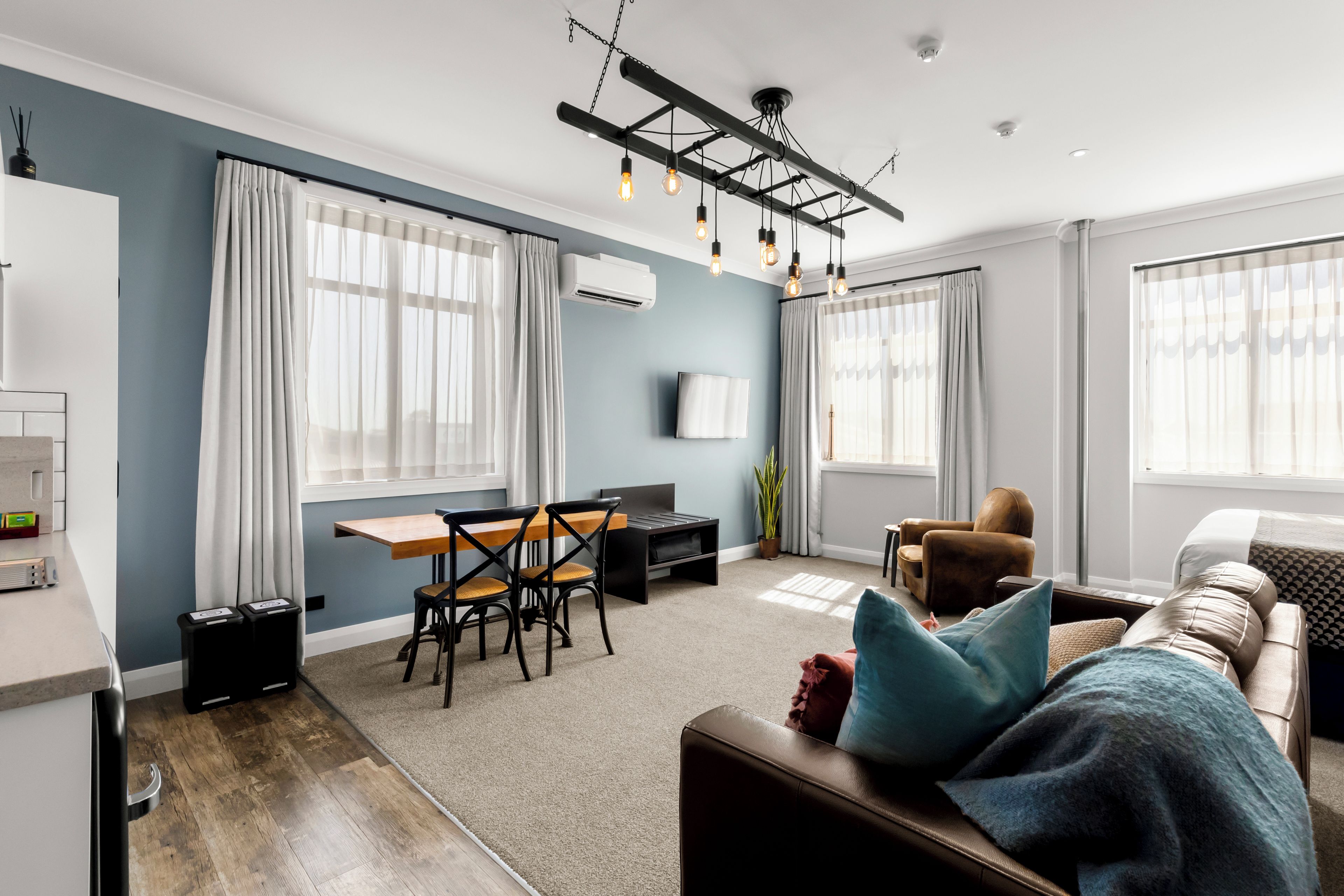 A modern living room with a dining area, a sofa with cushions, a TV mounted on the wall, and an air conditioning unit.