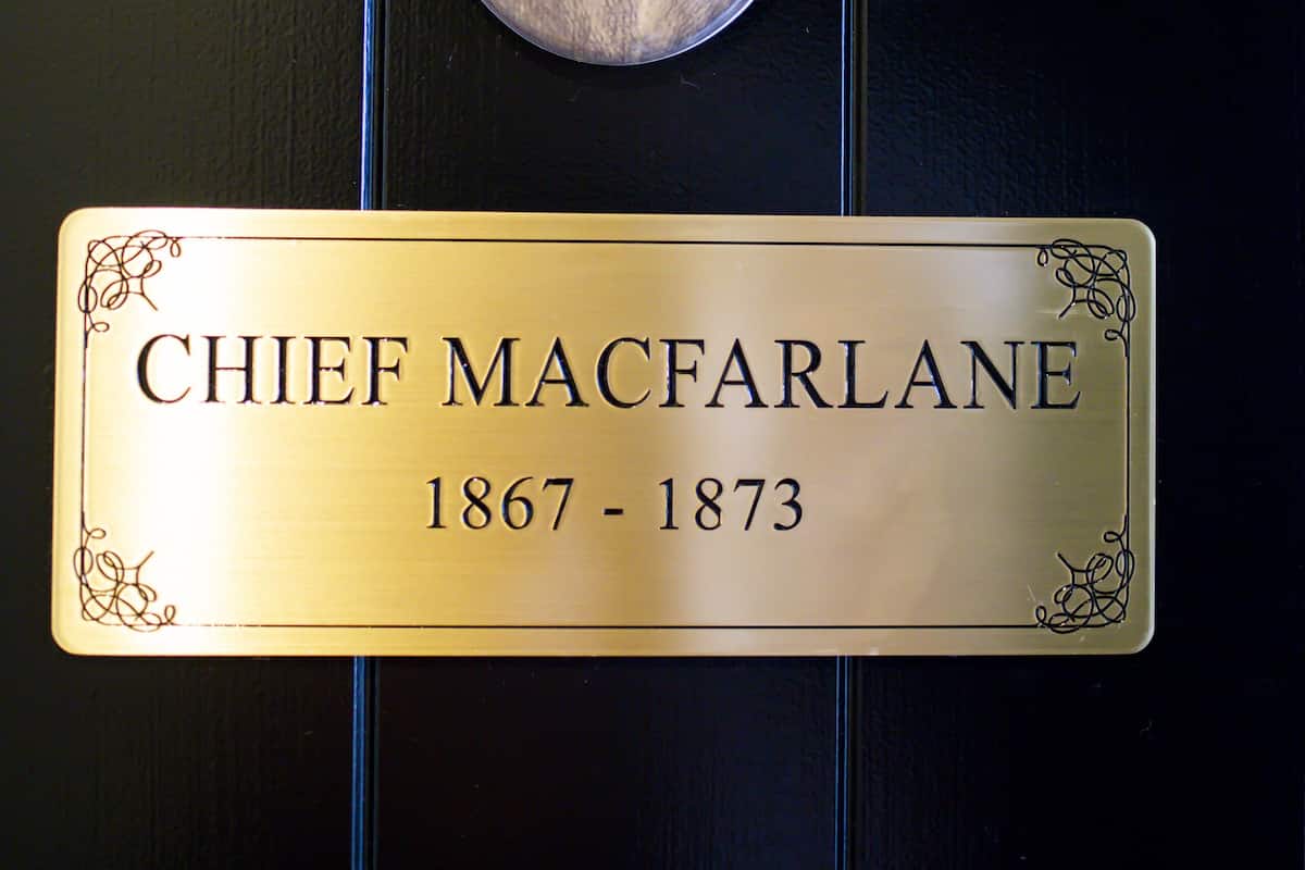 Eine Tafel mit dem Namen "Chief MacFarlane" und den Jahreszahlen "1867 - 1873" an einer dunklen Holztür angebracht.