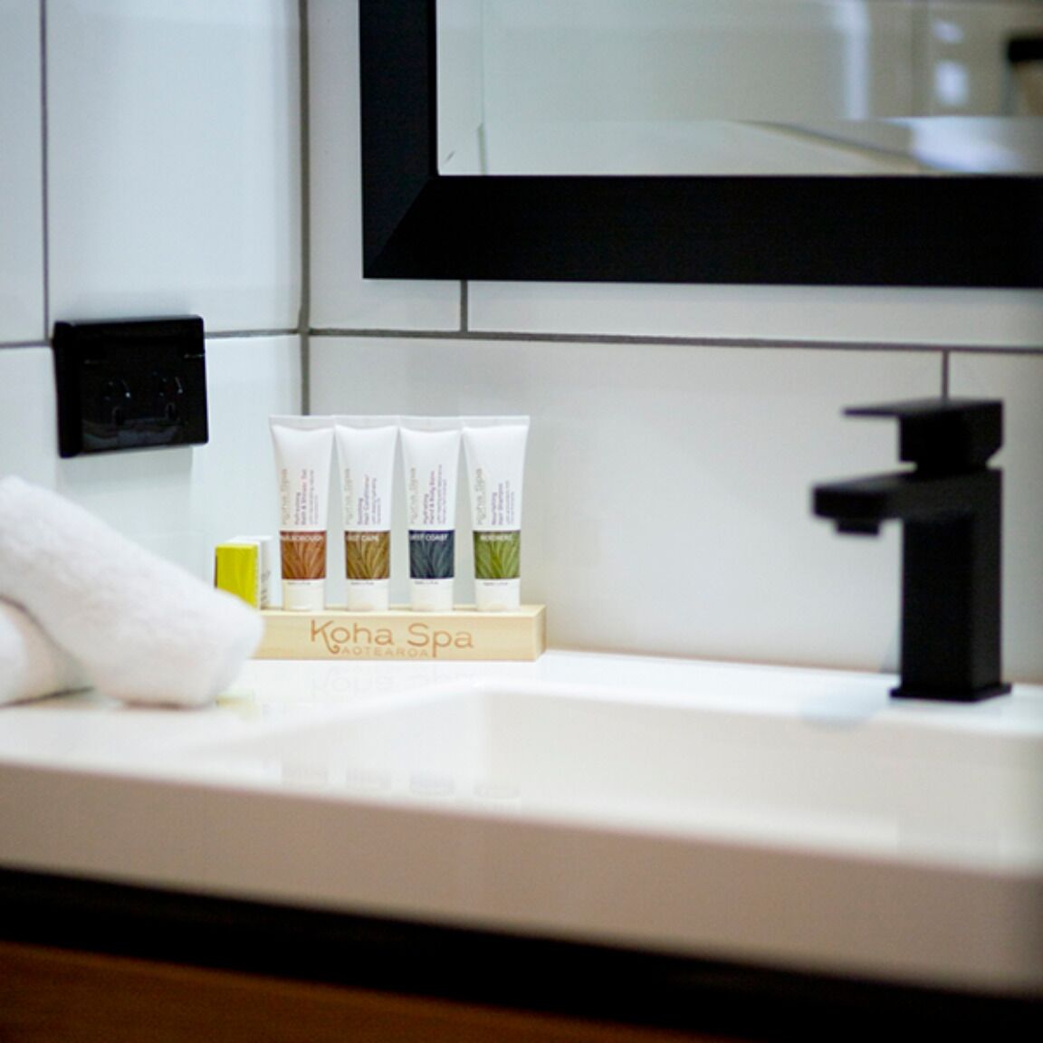 A bathroom counter with a white sink, a black faucet, and a towel. There are four tubes of lotion on a wooden stand labeled "Koha Spa."