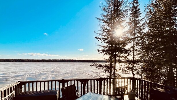 A deck with a table and chairs overlooking a frozen lake, surrounded by tall trees and a clear blue sky. - Cozy cottage in paradise! Cross Lake, Aroostook County (Cross Lake Township)