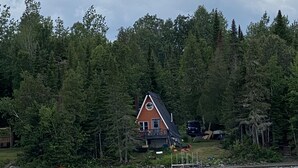 A cabin with a sloped roof and a deck, surrounded by dense forest and a body of water. - Cozy cottage in paradise! Cross Lake, Aroostook County (Cross Lake Township)