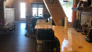 A cozy, rustic kitchen with wooden walls and a sloped ceiling. A dining table with chairs is positioned near a window, and various kitchen appliances and utensils are visible. - Cozy cottage in paradise! Cross Lake, Aroostook County (Cross Lake Township)