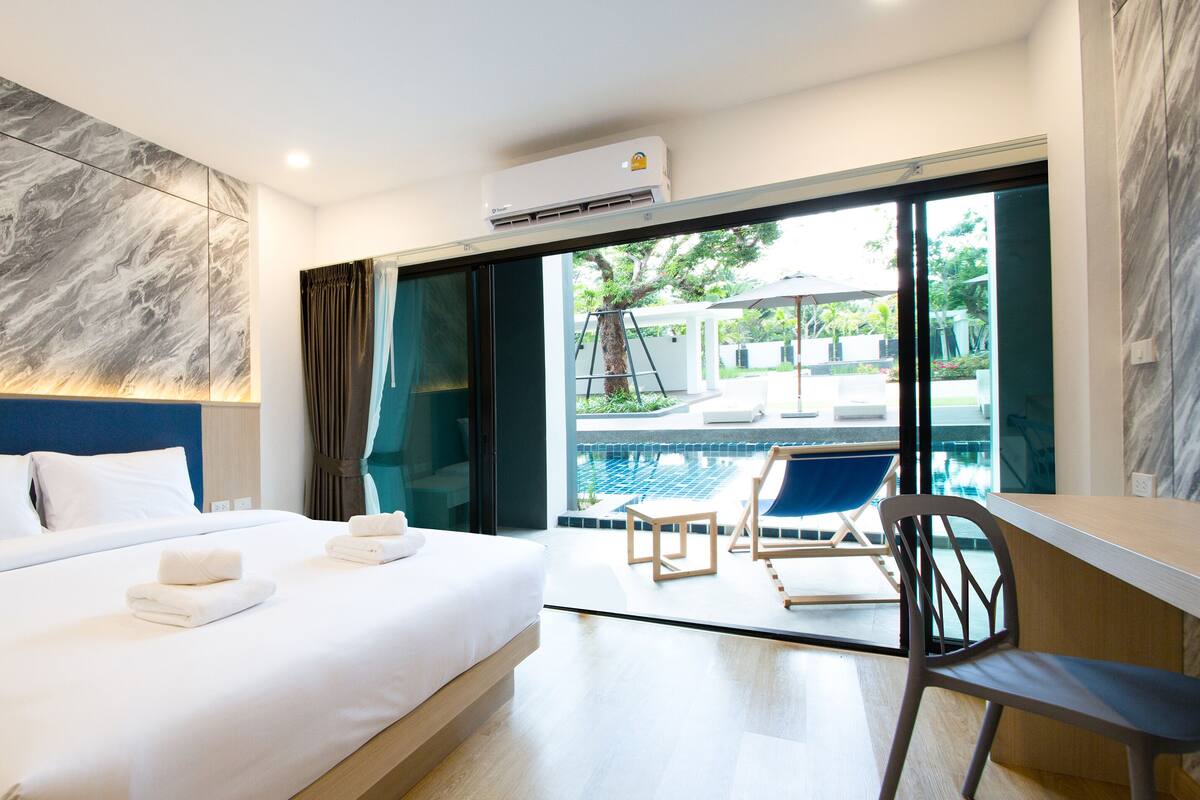Una habitación de hotel moderna con una cama grande, un vidrio que da a un área de piscina y un equipo de aire acondicionado en el techo.