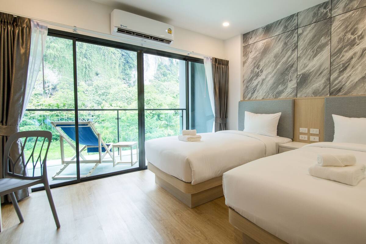 Habitación de hotel con dos camas, un balcón con una silla y vistas a zonas verdes.