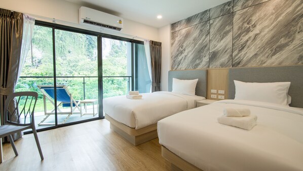 A hotel room with two beds, a balcony with a chair, and a view of greenery.