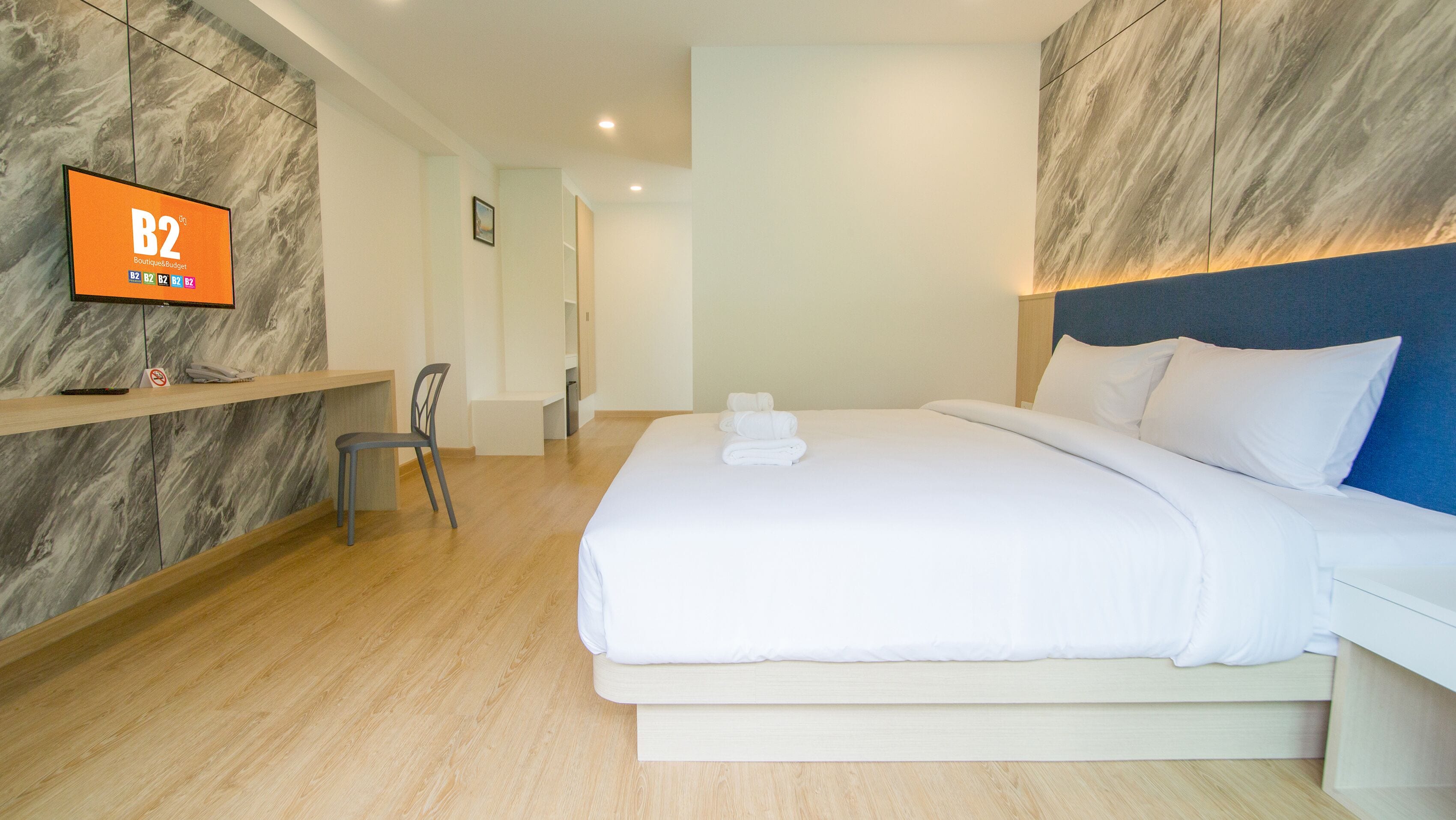 A hotel room with a large bed, a TV on the wall, a desk with a chair, and a bathroom in the background.