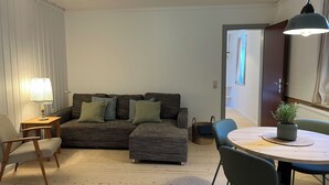 Una sala de estar acogedora con un sofá, una mesita pequeña y una silla. Hay una lámpara en una mesita auxiliar y una planta en maceta sobre una mesa.