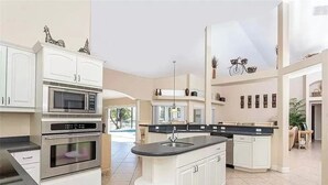 Una cocina moderna con armarios blancos, una isla central, electrodomésticos de acero inoxidable y vistas al exterior.