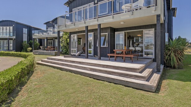 A modern two-story house with a balcony, a patio with a table and chairs, and a well-maintained lawn. - Golfer's Paradise - Matarangi Holiday Home (Matarangi)