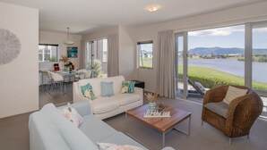 A modern living room with a sofa, a coffee table, and a wicker chair. There is a dining area with a table and chairs, and a view of a lake and mountains through large windows. - Golfer's Paradise - Matarangi Holiday Home (Matarangi)