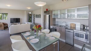 A modern kitchen with stainless steel appliances, a dining area with a glass table, and a living area with a sofa and TV. - Golfer's Paradise - Matarangi Holiday Home (Matarangi)